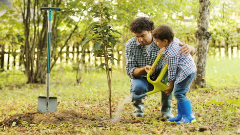Portrait-of-a-boy-and-his-dad-planting-a-tree.-They-water-the-tree.-Dad-helps-his-son.-Blurred-background